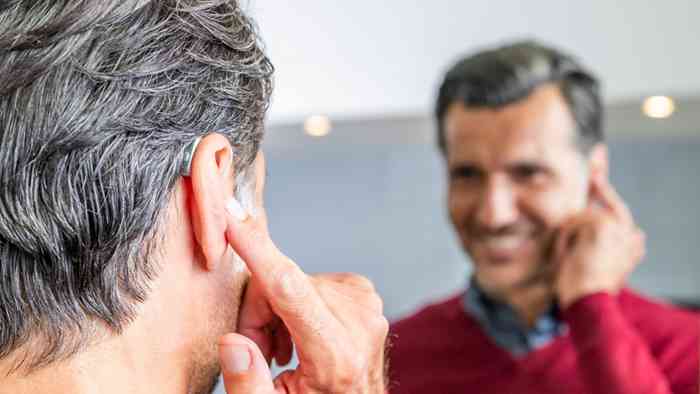 A man looking in the mirror while putting a hearing aid in his right ear